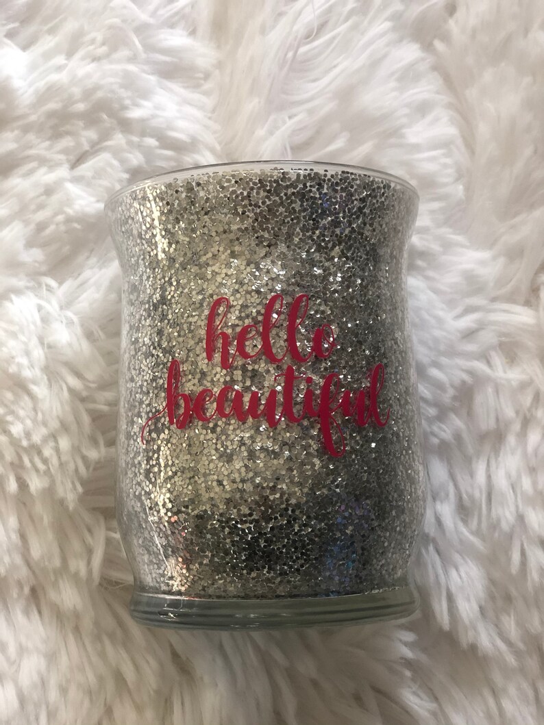 May include: A clear glass jar filled with silver glitter. The words "hello beautiful" are written in pink script across the front of the jar. The jar sits on a fluffy white surface.