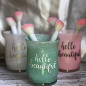 May include: Three glittery cosmetic brush holders in green, pink, and white, each with the words "hello beautiful" in cursive. The holders contain makeup brushes with white handles and pink brush heads. The background is blurred.