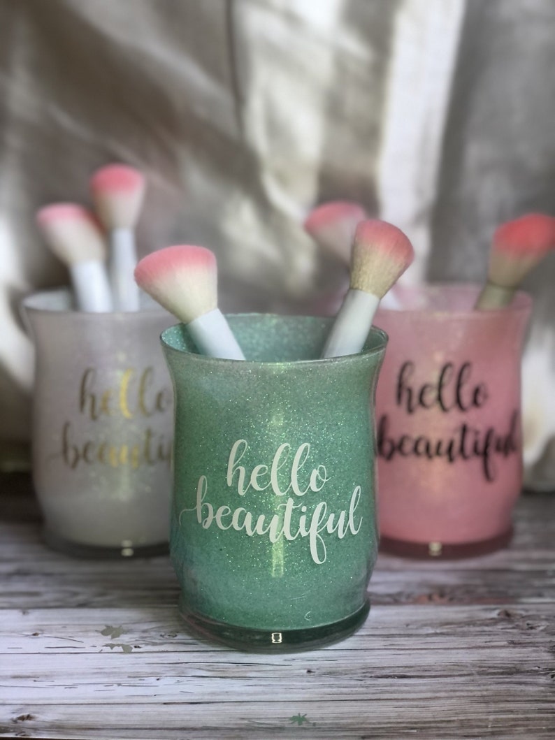 May include: Three glittery cosmetic brush holders in green, pink, and white, each with the words "hello beautiful" in cursive. The holders contain makeup brushes with white handles and pink brush heads. The background is blurred.