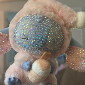 May include: A plush keychain featuring a character with a light pink fuzzy hood and body, blue hands and feet, and a face covered in iridescent rhinestones. The character holds a small tan and white drink cup with a straw, attached to a metal ring.