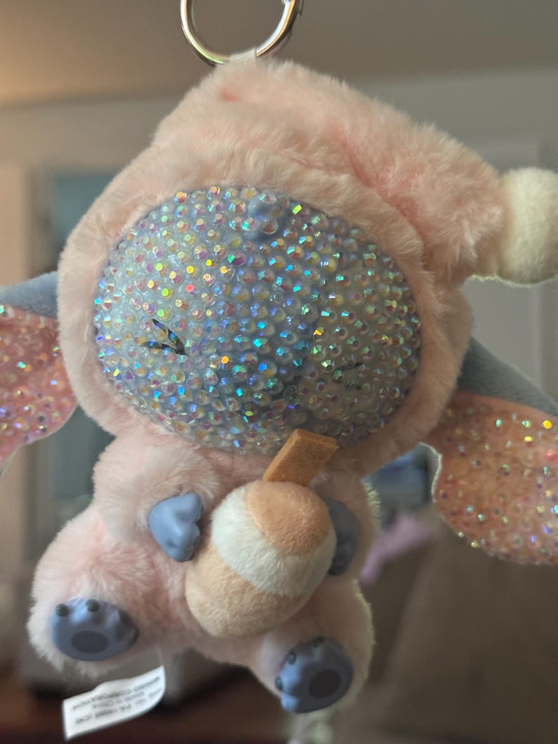 May include: A plush keychain featuring a character with a light pink fuzzy hood and body, blue hands and feet, and a face covered in iridescent rhinestones. The character holds a small tan and white drink cup with a straw, attached to a metal ring.