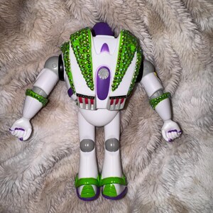 Pode incluir: Um brinquedo rob&ocirc; branco e verde com detalhes em roxo. O rob&ocirc; tem pedras preciosas verdes nas costas e nos bra&ccedil;os. Os p&eacute;s do rob&ocirc; s&atilde;o verdes e roxos. Os bra&ccedil;os do rob&ocirc; est&atilde;o estendidos. A palavra "DANGER" &eacute; vis&iacute;vel nas costas do rob&ocirc;.