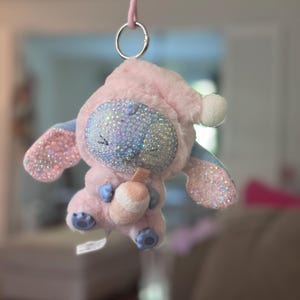 May include: A plush character ornament in a light pink hooded outfit, featuring a face covered in iridescent rhinestones. The character has large ears with pink glitter accents, holds a small drink cup, and hangs from a pink cord with a metal ring.