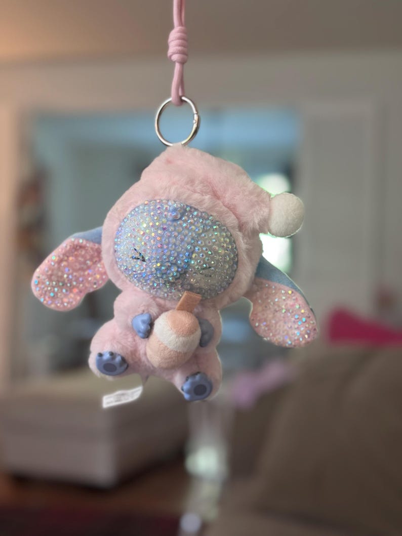 May include: A plush character ornament in a light pink hooded outfit, featuring a face covered in iridescent rhinestones. The character has large ears with pink glitter accents, holds a small drink cup, and hangs from a pink cord with a metal ring.