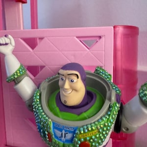 Pode incluir: Figura de a&ccedil;&atilde;o do Buzz Lightyear com um traje espacial verde e branco, cabe&ccedil;a roxa e express&atilde;o sorridente. As palavras "SPACE RANGER" e "LIGHTYEAR" est&atilde;o no peito. O fundo &eacute; rosa.