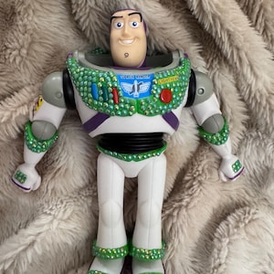 Pode incluir: Figura de a&ccedil;&atilde;o do Buzz Lightyear com um rosto sorridente, vestindo um traje espacial branco e verde. O traje espacial &eacute; adornado com strass verdes e vermelhos. As palavras "SPACE RANGER" e "LIGHTYEAR" s&atilde;o vis&iacute;veis no peito.