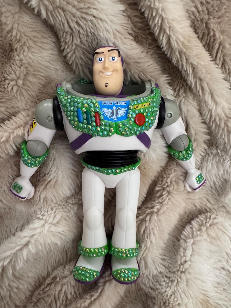 Pode incluir: Figura de a&ccedil;&atilde;o do Buzz Lightyear com um rosto sorridente, vestindo um traje espacial branco e verde. O traje espacial &eacute; adornado com strass verdes e vermelhos. As palavras "SPACE RANGER" e "LIGHTYEAR" s&atilde;o vis&iacute;veis no peito.