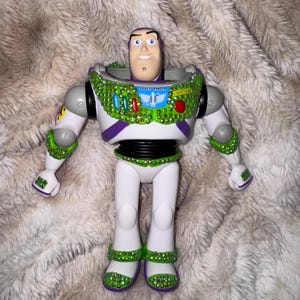 Pode incluir: Figura de a&ccedil;&atilde;o Buzz Lightyear com corpo branco, detalhes verdes e roxos. A figura &eacute; adornada com enfeites verdes e vermelhos brilhantes ao redor da gola, bra&ccedil;os e pernas. As palavras "SPACE RANGER" e "LIGHTYEAR" s&atilde;o vis&iacute;veis.