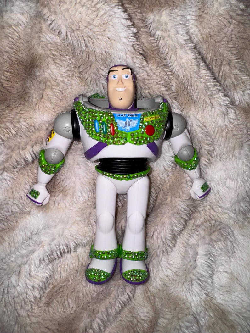 Pode incluir: Figura de a&ccedil;&atilde;o Buzz Lightyear com corpo branco, detalhes verdes e roxos. A figura &eacute; adornada com enfeites verdes e vermelhos brilhantes ao redor da gola, bra&ccedil;os e pernas. As palavras "SPACE RANGER" e "LIGHTYEAR" s&atilde;o vis&iacute;veis.