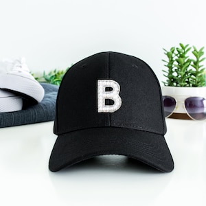 May include: Black baseball cap with a rhinestone letter "B" on the front. The cap is displayed on a white surface with white sneakers, sunglasses, and a water bottle in the background. The cap is a stylish accessory.