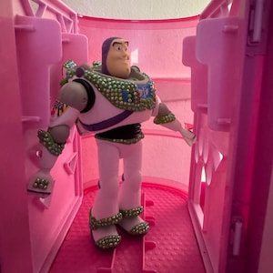 Pode incluir: Uma figura de a&ccedil;&atilde;o do Buzz Lightyear est&aacute; dentro de um compartimento de pl&aacute;stico rosa. A figura est&aacute; vestida com um traje espacial branco e verde com um cinto roxo e preto. O brinquedo tem um capacete verde e branco e uma express&atilde;o determinada.