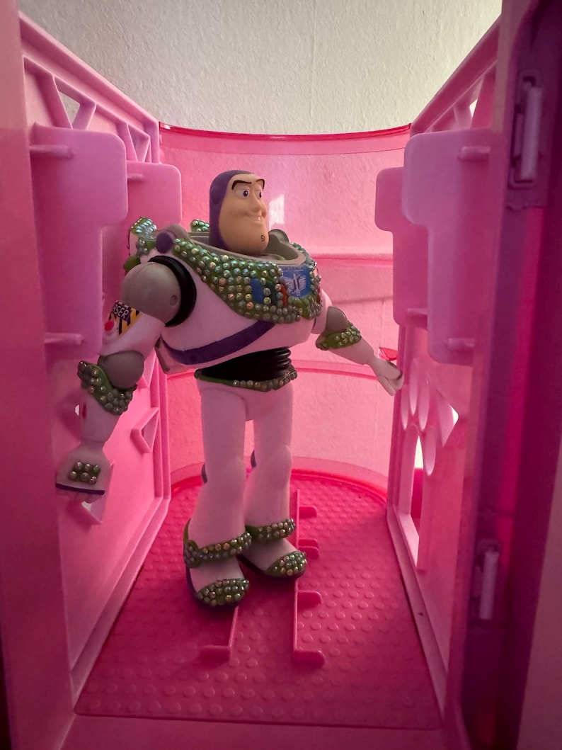 Pode incluir: Uma figura de a&ccedil;&atilde;o do Buzz Lightyear est&aacute; dentro de um compartimento de pl&aacute;stico rosa. A figura est&aacute; vestida com um traje espacial branco e verde com um cinto roxo e preto. O brinquedo tem um capacete verde e branco e uma express&atilde;o determinada.