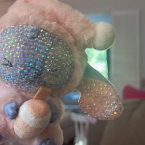 May include: A plush keychain featuring a light pink character with a soft, fuzzy texture. The character has a large, rounded face covered in iridescent, light-reflecting rhinestones, a small tan drink accessory, and light blue paws and ears.