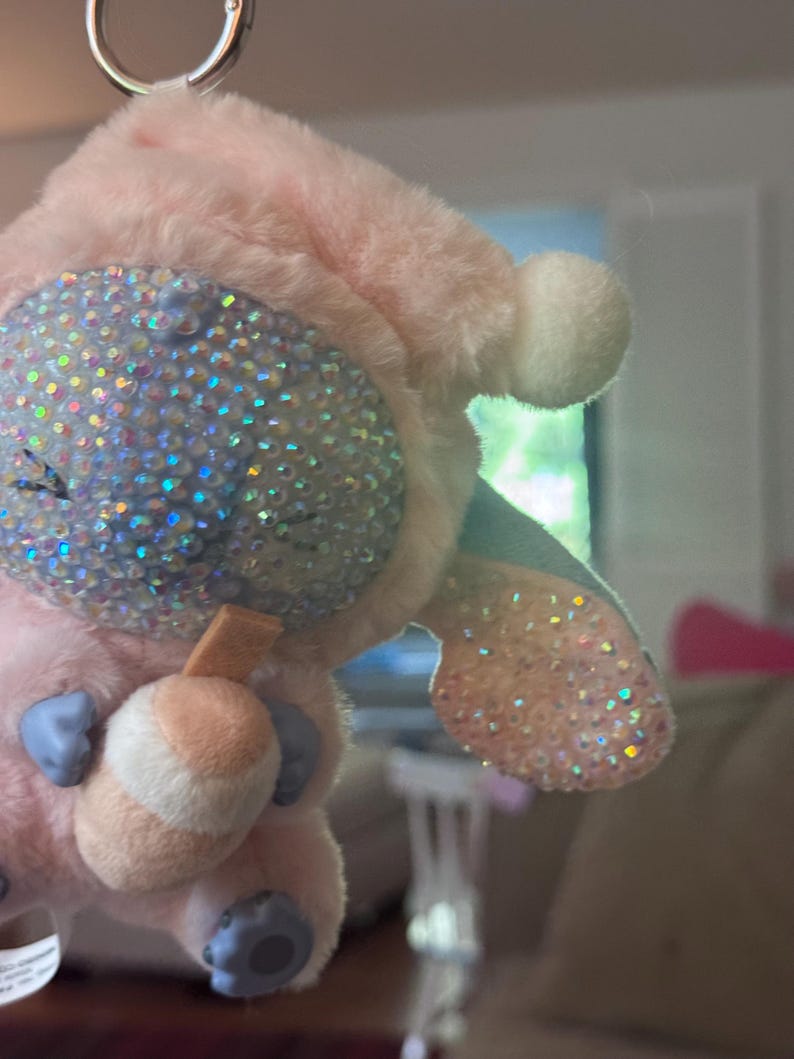 May include: A plush keychain featuring a light pink character with a soft, fuzzy texture. The character has a large, rounded face covered in iridescent, light-reflecting rhinestones, a small tan drink accessory, and light blue paws and ears.