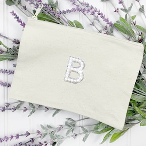 May include: A cream-colored canvas zippered pouch with a large, embellished letter "B" in the center. The letter is decorated with iridescent and silver-toned beads. The pouch is set against a backdrop of lavender sprigs.