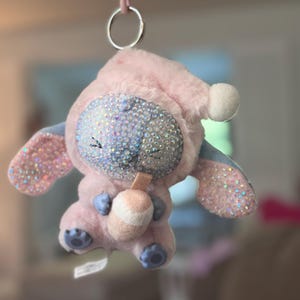 May include: A plush character keychain featuring a light pink hooded outfit, a face covered in iridescent rhinestones, and large ears with rhinestone accents. The character holds a small, light-colored drink cup and hangs from a silver metal ring.