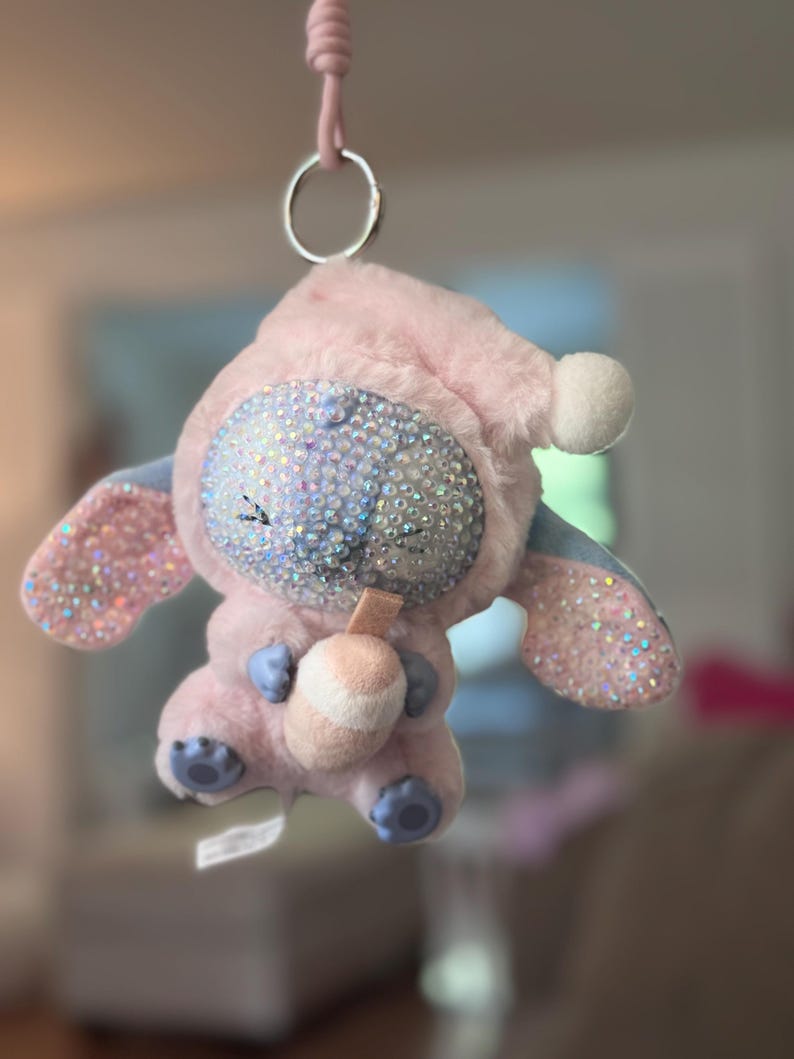 May include: A plush character keychain featuring a light pink hooded outfit, a face covered in iridescent rhinestones, and large ears with rhinestone accents. The character holds a small, light-colored drink cup and hangs from a silver metal ring.
