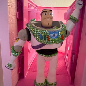 Pode incluir: Figura de a&ccedil;&atilde;o do Buzz Lightyear, com um traje espacial branco e verde com um detalhe roxo. A figura tem um rosto sorridente e est&aacute; posando com um bra&ccedil;o levantado. A figura &eacute; decorada com strass verdes e transparentes. As palavras "SPACE RANGER" s&atilde;o vis&iacute;veis.