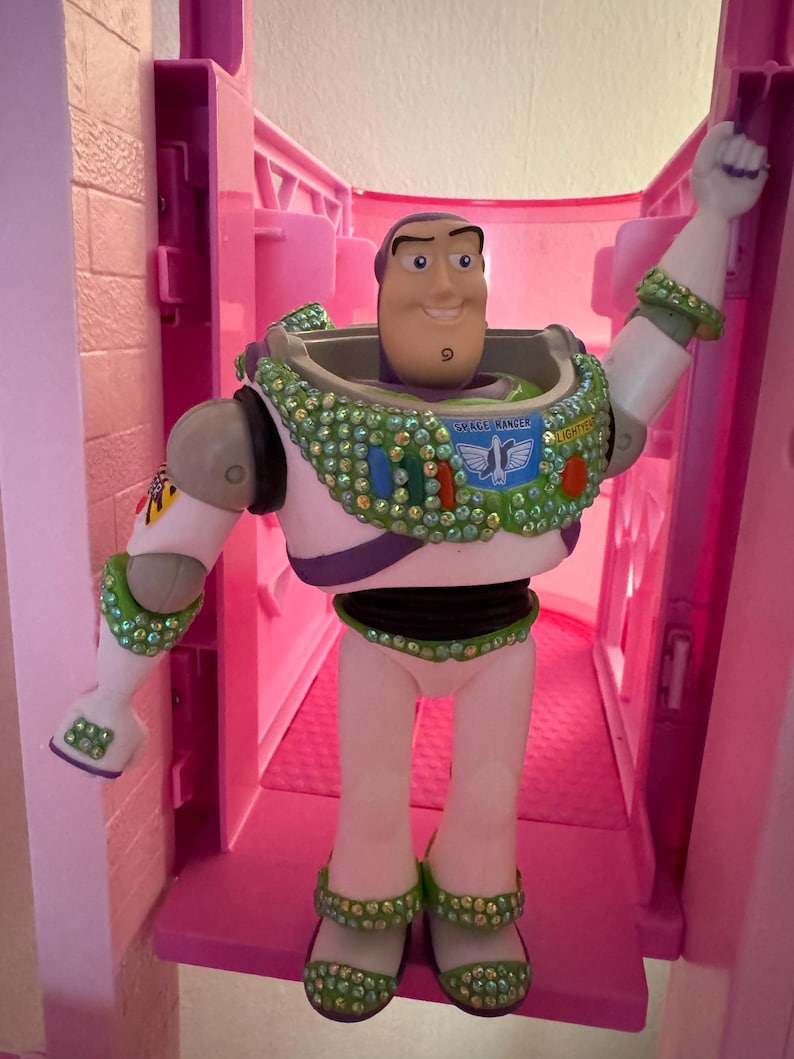 Pode incluir: Figura de a&ccedil;&atilde;o do Buzz Lightyear, com um traje espacial branco e verde com um detalhe roxo. A figura tem um rosto sorridente e est&aacute; posando com um bra&ccedil;o levantado. A figura &eacute; decorada com strass verdes e transparentes. As palavras "SPACE RANGER" s&atilde;o vis&iacute;veis.