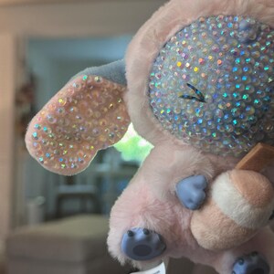 May include: A plush character toy featuring soft pink fur, light blue accents on the feet and hands, and a face and ears covered in iridescent, multi-colored rhinestones. The character holds a small, light-colored cylindrical object in its hands.