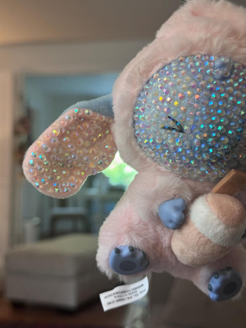 May include: A plush character toy featuring soft pink fur, light blue accents on the feet and hands, and a face and ears covered in iridescent, multi-colored rhinestones. The character holds a small, light-colored cylindrical object in its hands.