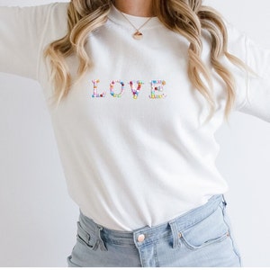 Pode incluir: Camisola branca com a palavra "LOVE" bordada no peito em letras coloridas de contas. As letras são uma mistura de contas rosa, azul, amarelo e verde. A camisola tem mangas compridas e gola redonda.