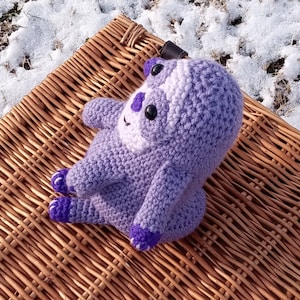 May include: A purple crocheted sloth toy sitting on a wicker basket. The sloth has a white face and black eyes.