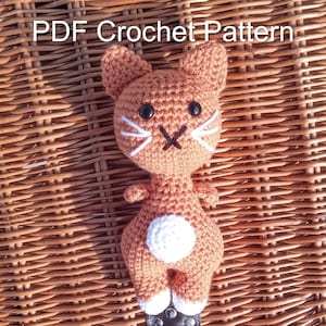May include: Crocheted orange cat amigurumi toy with white accents. The cat has black eyes and a white nose. The toy is sitting on a wicker surface. PDF Crochet Pattern.