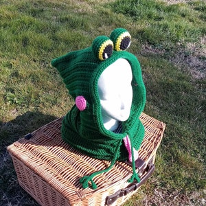 May include: Green crocheted frog hooded scarf with pink and yellow accents. The scarf is sitting on a wicker basket.