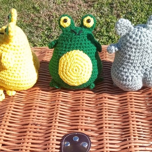 May include: Three crocheted stuffed animals: a yellow and green frog, a green frog with a yellow belly, and a gray elephant. The animals are sitting on a wicker basket.