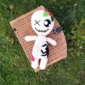 May include: White crocheted doll with black and red details. The doll has a stitched-on smile and cross-shaped eyes. The doll is lying on a wicker basket.