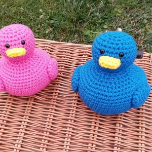 Op de afbeelding: Twee gehaakte eendjes, een roze en een blauwe, met gele snavels en zwarte ogen. De eendjes zitten op een geweven rieten oppervlak.