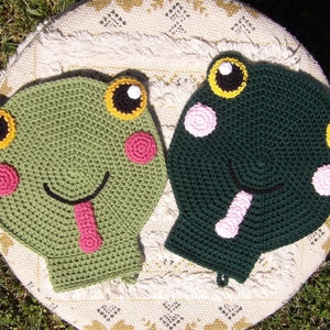 Puede incluir: Dos agarraderas de olla de ganchillo con forma de rana. Una es verde con detalles en rosa y amarillo, la otra es verde oscuro con detalles en rosa y amarillo.