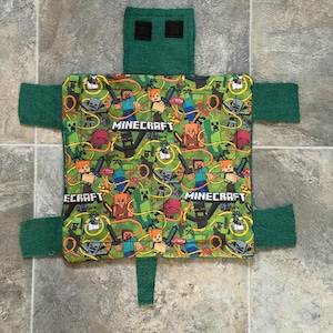 Könnte beinhalten: Eine grüne und gemusterte Babydecke im Minecraft-Design. Die Decke hat ein zentrales Quadrat mit einem sich wiederholenden Muster von Minecraft-Charakteren und -Logos. Kopf und Gliedmaßen sind aus einfarbigem grünem Stoff.