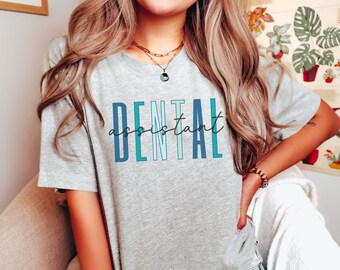 Op de afbeelding: Een grijs T-shirt met de tekst "DENTAL assistant" in blauwe, groene en witte letters.