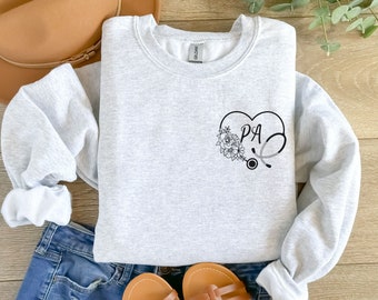 May include: Gray sweatshirt with a black heart design featuring the letters "PA" and a stethoscope. The heart is surrounded by floral details.
