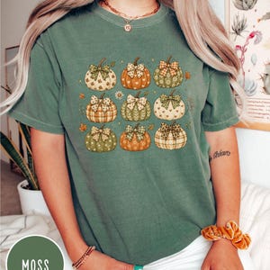 May include: A moss green short-sleeve t-shirt featuring a graphic print of nine pumpkins arranged in a grid. Each pumpkin displays a unique pattern, such as plaid, hearts, or floral motifs, and is topped with a decorative bow in various colors.