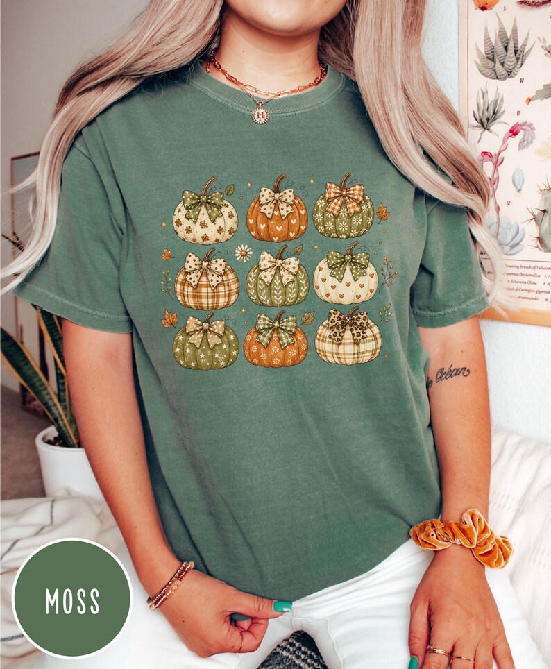 May include: A moss green short-sleeve t-shirt featuring a graphic print of nine pumpkins arranged in a grid. Each pumpkin displays a unique pattern, such as plaid, hearts, or floral motifs, and is topped with a decorative bow in various colors.