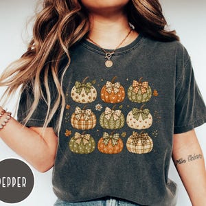 May include: A charcoal gray short-sleeve t-shirt featuring a graphic print of nine pumpkins arranged in a grid. Each pumpkin displays a unique pattern, such as plaid, hearts, or floral designs, and is topped with a decorative bow in coordinating colors.