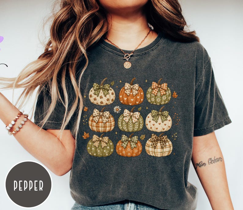 May include: A charcoal gray short-sleeve t-shirt featuring a graphic print of nine pumpkins arranged in a grid. Each pumpkin displays a unique pattern, such as plaid, hearts, or floral designs, and is topped with a decorative bow in coordinating colors.