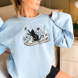 May include: A light blue sweatshirt with a black cat sitting on an open book with flowers and a butterfly. The cat is looking at the butterfly.