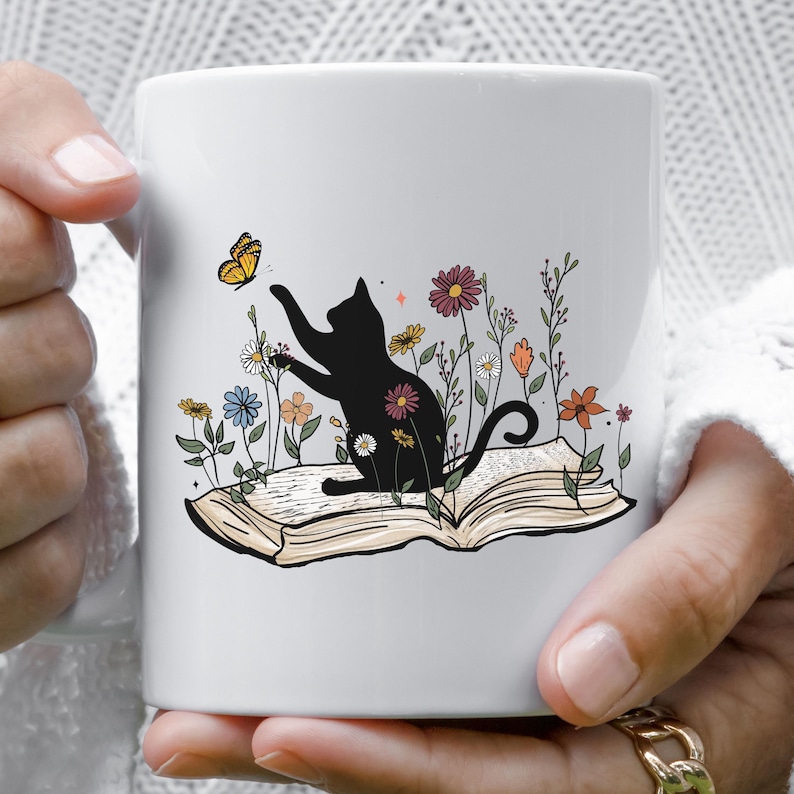 May include: White ceramic mug with a black cat sitting on an open book. The cat is reaching for a yellow butterfly. The book is surrounded by colorful flowers.