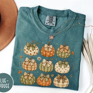 May include: A blue-spruce colored short-sleeve t-shirt featuring a graphic print of nine pumpkins in various shades of orange, green, and cream. Each pumpkin is decorated with a patterned bow and small stars, hearts, or floral motifs.