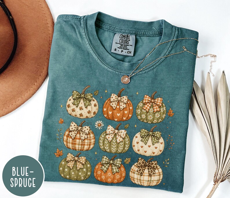 May include: A blue-spruce colored short-sleeve t-shirt featuring a graphic print of nine pumpkins in various shades of orange, green, and cream. Each pumpkin is decorated with a patterned bow and small stars, hearts, or floral motifs.
