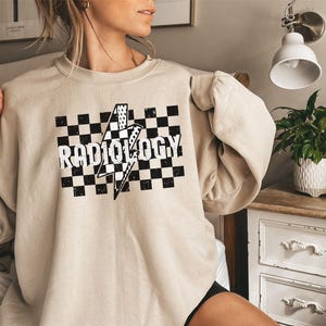 May include: A beige sweatshirt with a black and white checkered pattern and the word "RADIOLOGY" in black text. A lightning bolt graphic is centered in the checkered pattern.