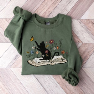 May include: A green sweatshirt with a black cat sitting on an open book. The cat is reaching for a yellow butterfly. There are flowers around the book and cat.