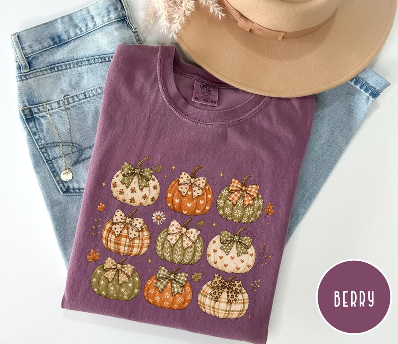 May include: A berry-colored crew neck t-shirt featuring a graphic print of nine pumpkins, each decorated with a unique patterned bow. The pumpkins display various colors and textures, including plaid, polka dots, and floral motifs, arranged on the front.