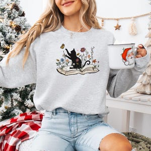 May include: A gray sweatshirt with a black cat silhouette sitting on an open book with flowers and a butterfly. The cat is reaching for the butterfly.