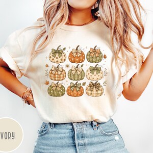 May include: An ivory t-shirt featuring a graphic print of nine pumpkins arranged in a grid. Each pumpkin displays a unique pattern, including plaid, polka dots, and hearts, and is topped with a decorative bow. Small autumn leaves accent the design.