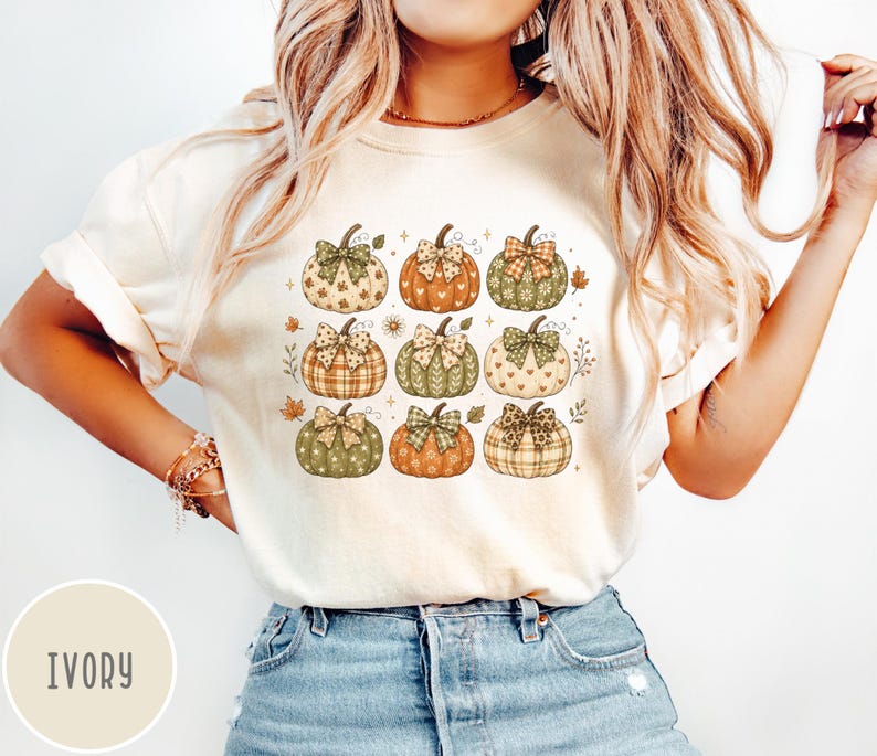 May include: An ivory t-shirt featuring a graphic print of nine pumpkins arranged in a grid. Each pumpkin displays a unique pattern, including plaid, polka dots, and hearts, and is topped with a decorative bow. Small autumn leaves accent the design.